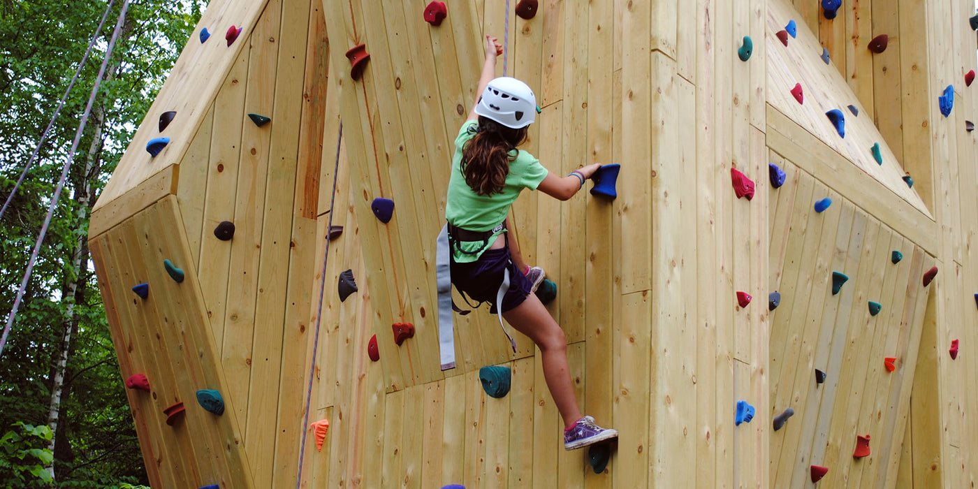 Teenage girl, wearing a white safety helmet and black harness, climbs an outdoor climbing tower designed and built by ABEE Inc. in Bemidji, Minnesota.