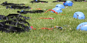 Black challenge course harnesses and blue helmets lined up and laying on the green grass.