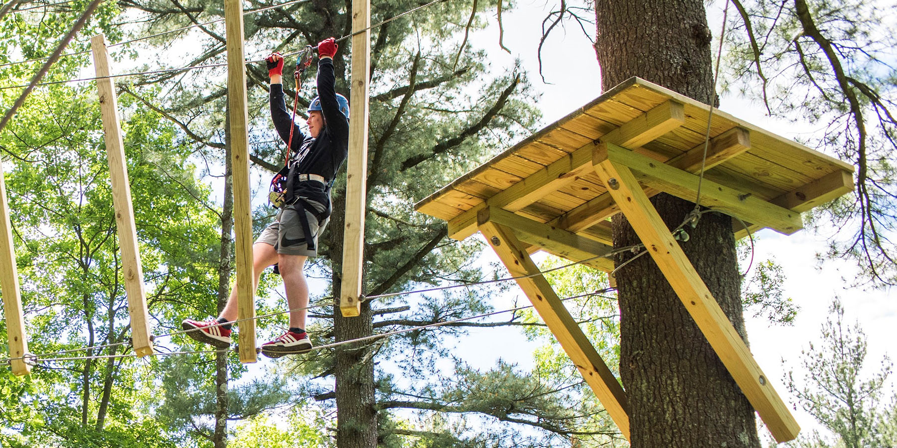 Young adult in a safety harness walking a rope bridge on a challenge course designed and built by ABEE Inc. in Wisconsin Dells.