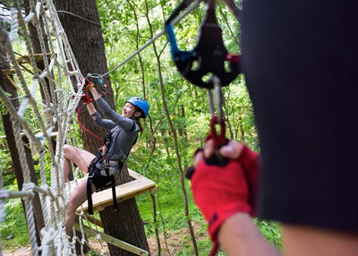 A young woman in a black climbing harness attached to a wire cable and wearing a blue climbing helmet, safely traverses a rope net attached to a canopy challenge course designed and built by ABEE Inc. in Wisconsin Dells, Wisconsin.